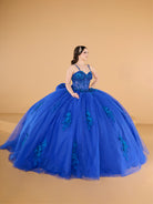 A woman modeling a voluminous royal blue ball gown with floral lace applique and a sweetheart neckline, posed against a solid beige studio backdrop. / This ball gown features a fitted, embellished bodice with thin straps and a full skirt decorated with floral appliqué, finished with a bow at the back.
