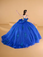 Woman in a blue ball gown with floral details on a beige background / This ball gown features a fitted, embellished bodice with thin straps and a full skirt decorated with floral appliqué, finished with a bow at the back.