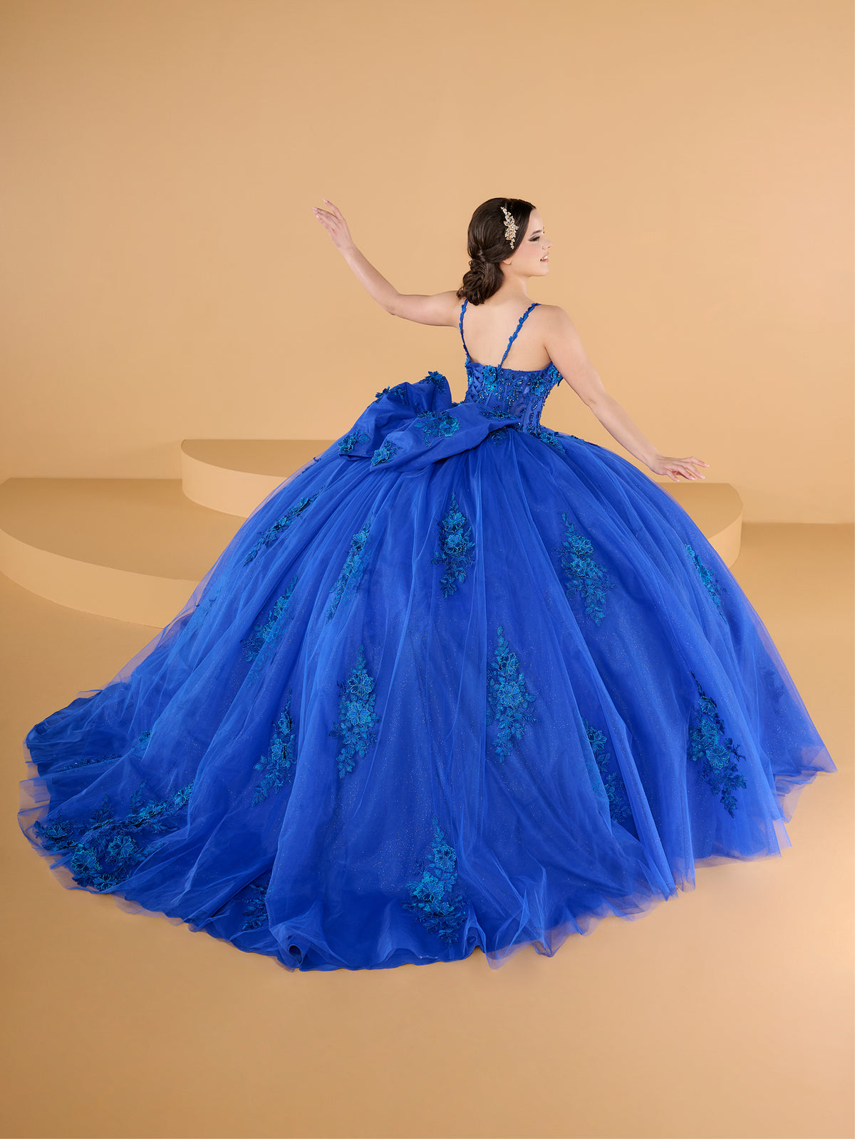 Woman in a blue ball gown with floral details on a beige background / This ball gown features a fitted, embellished bodice with thin straps and a full skirt decorated with floral appliqué, finished with a bow at the back.