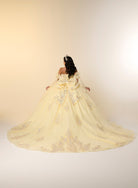 A woman viewed from behind wearing an off-the-shoulder ball gown with a very full skirt and long train in pale yellow with silver floral embroidery, set against a warm beige studio background. / This elegant ball gown features a strapless straight neckline with delicate draped sleeves. The full skirt is crafted from soft tulle and adorned with shimmering embroidery, while a large bow at the back adds a graceful finishing touch.