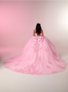 A woman viewed from behind wearing a voluminous light pink ball gown with a large bow at the back, set against a soft pink and white studio backdrop. / This elegant ball gown features a strapless straight neckline with delicate draped sleeves. The full skirt is crafted from soft tulle and adorned with shimmering embroidery, while a large bow at the back adds a graceful finishing touch.