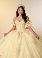 A woman wearing an elaborate off-the-shoulder yellow quinceañera dress with intricate beadwork, a matching tiara, and a necklace, posed against a solid beige studio background. / This elegant ball gown features a strapless straight neckline with delicate draped sleeves. The full skirt is crafted from soft tulle and adorned with shimmering embroidery, while a large bow at the back adds a graceful finishing touch.