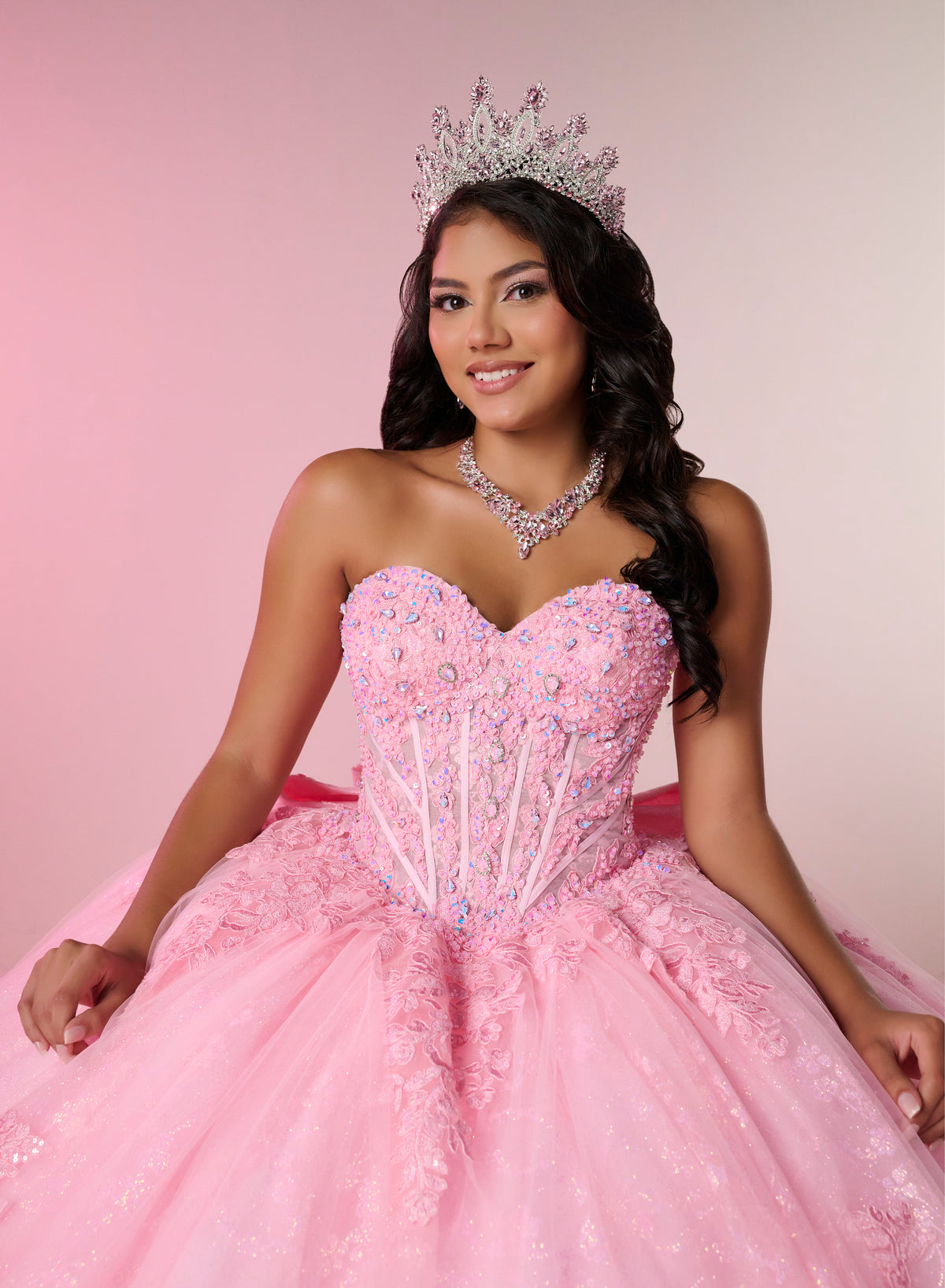 A woman wearing a sparkling pink quinceañera ball gown with a strapless sweetheart bodice, beaded corset-style waist, and full tulle skirt, seated against a soft pink studio background. She is also wearing a silver tiara and matching silver necklace. / This elegant ball gown features a strapless straight neckline with delicate draped sleeves. The full skirt is crafted from soft tulle and adorned with shimmering embroidery, while a large bow at the back adds a graceful finishing touch.