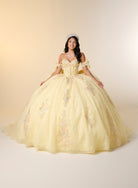 A woman wearing a voluminous pale yellow ball gown with off-the-shoulder straps, a fitted embellished bodice, and embroidered tulle details, posed against a warm cream studio background. / This elegant ball gown features a strapless straight neckline with delicate draped sleeves. The full skirt is crafted from soft tulle and adorned with shimmering embroidery, while a large bow at the back adds a graceful finishing touch.