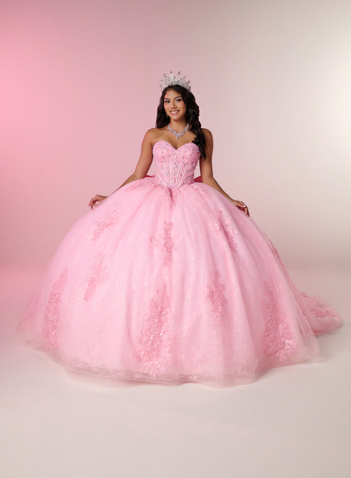 A young woman wearing a voluminous pink ball gown with a sweetheart bodice, lace applique, and a sparkling tiara, posed against a soft pink studio backdrop. / This elegant ball gown features a strapless straight neckline with delicate draped sleeves. The full skirt is crafted from soft tulle and adorned with shimmering embroidery, while a large bow at the back adds a graceful finishing touch.