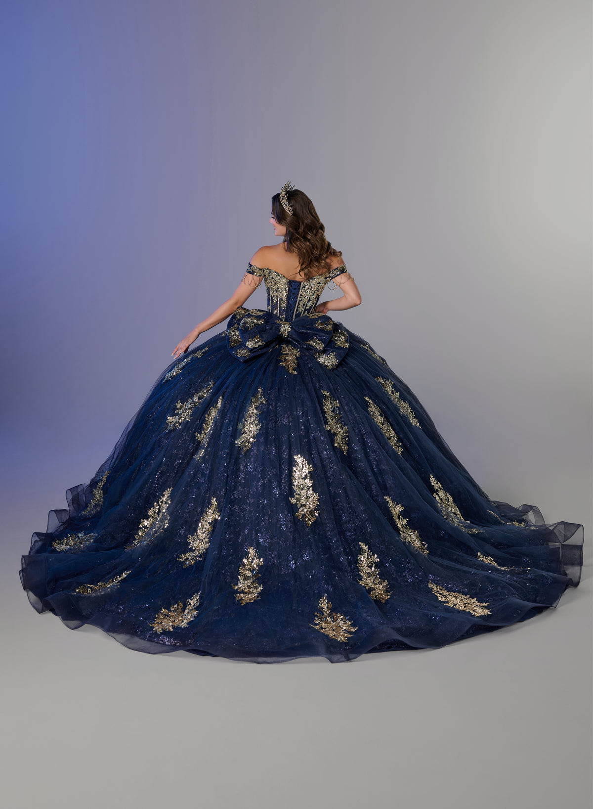 A model viewed from behind wearing a voluminous navy blue ball gown with intricate gold floral embroidery, set against a neutral gray studio background. / An off-the-shoulder ball gown featuring an embroidered bodice, a voluminous tulle skirt with floral applique panels, and a large accent bow on the back.