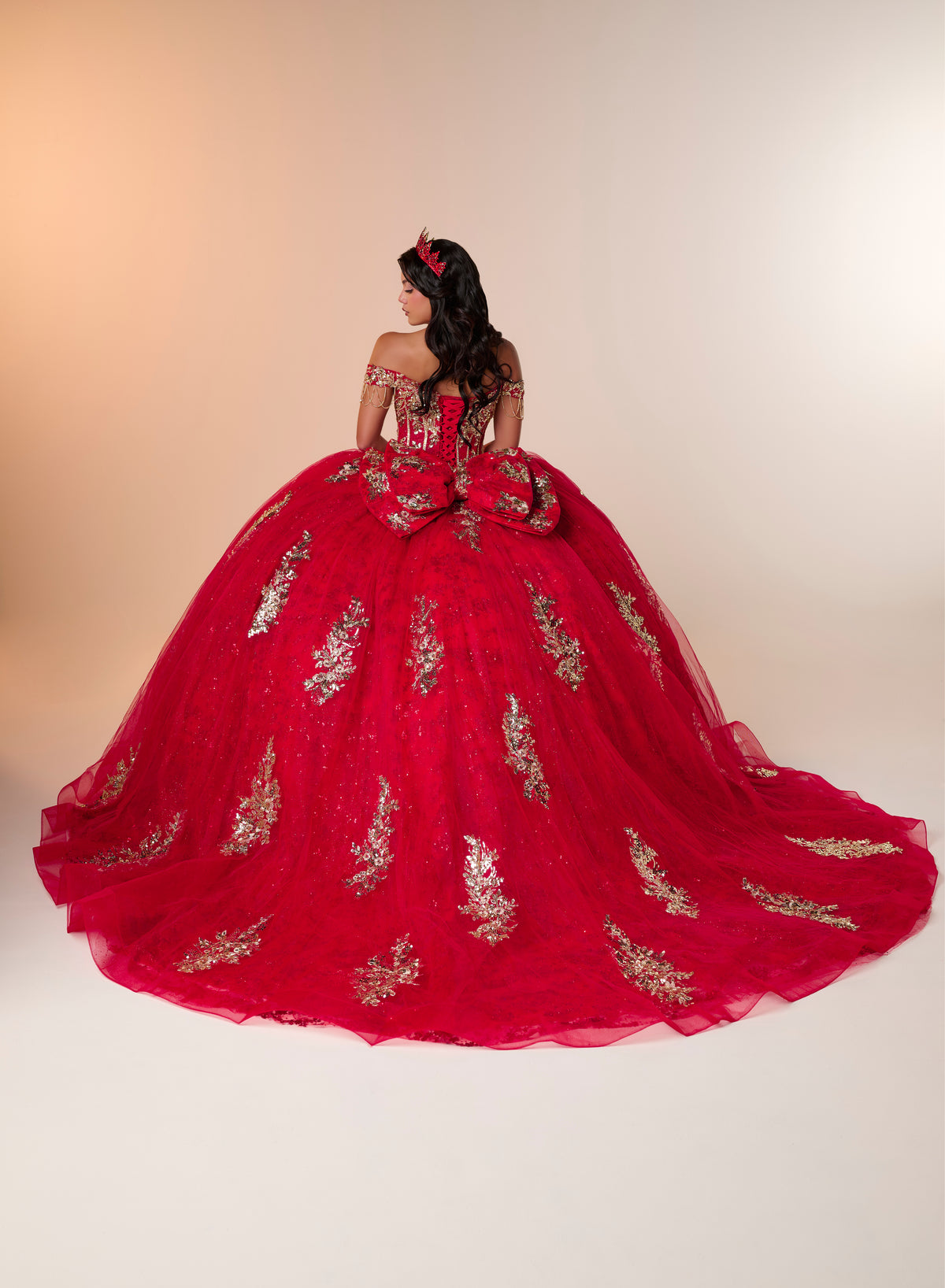 A woman wearing a voluminous red quinceañera ball gown with gold floral embroidery, photographed from behind against a warm beige studio backdrop. / An off-the-shoulder ball gown featuring an embroidered bodice, a voluminous tulle skirt with floral applique panels, and a large accent bow on the back.