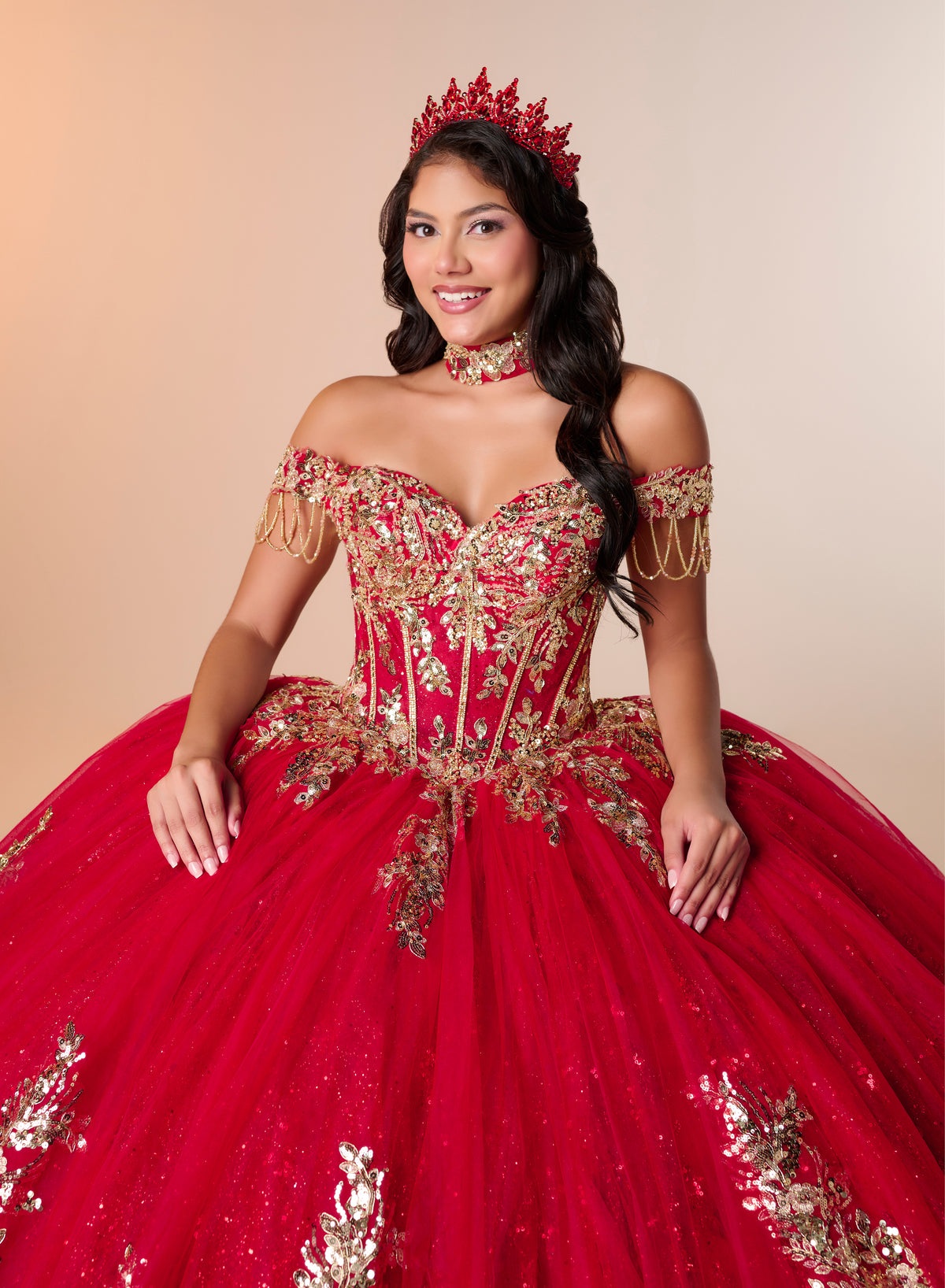A woman wearing a voluminous red quinceañera ball gown with gold embroidery, a matching red crown, and a red choker against a warm beige studio background. / An off-the-shoulder ball gown featuring an embroidered bodice, a voluminous tulle skirt with floral applique panels, and a large accent bow on the back.