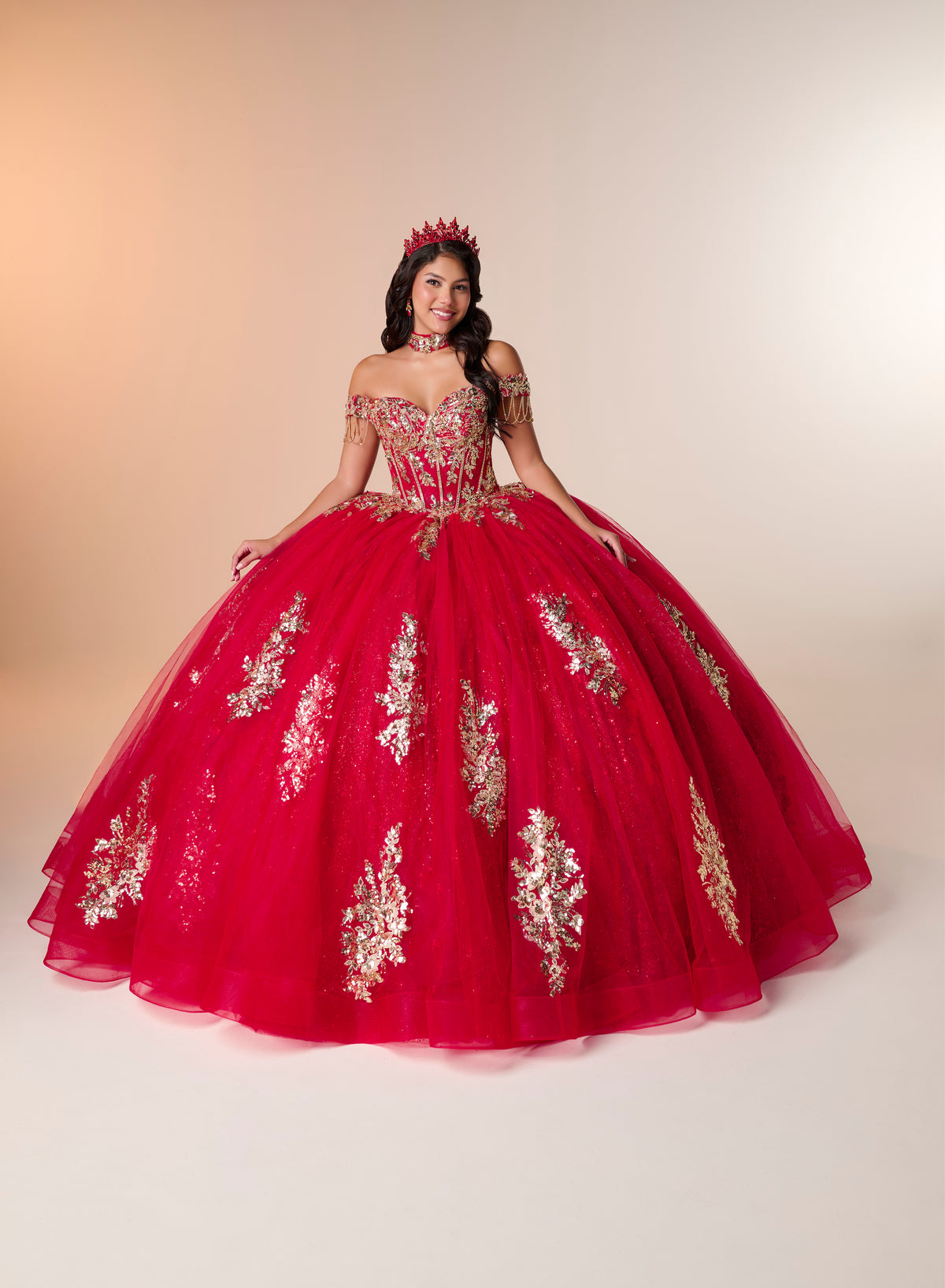 A woman modeling a voluminous red quinceañera ball gown with gold floral embroidery, a sweetheart neckline, and a matching red crown, set against a warm beige studio background. / An off-the-shoulder ball gown featuring an embroidered bodice, a voluminous tulle skirt with floral applique panels, and a large accent bow on the back.