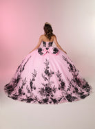 A young woman viewed from behind wearing a voluminous pink ball gown with black floral lace applique and a large bow at the back, posed against a soft pink-to-white studio backdrop. / A strapless ball gown with a lace-up bodice and a voluminous tulle skirt featuring floral embroidery and a large back bow.