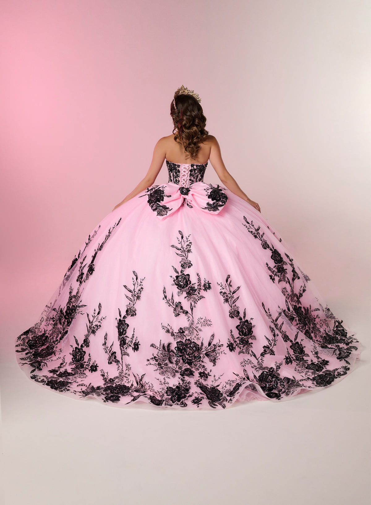 A young woman viewed from behind wearing a voluminous pink ball gown with black floral lace applique and a large bow at the back, posed against a soft pink-to-white studio backdrop. / A strapless ball gown with a lace-up bodice and a voluminous tulle skirt featuring floral embroidery and a large back bow.