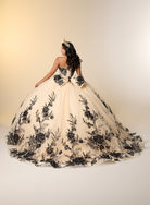 A woman wearing a strapless ball gown with black floral lace applique over a cream tulle base, photographed from the back against a warm beige studio backdrop. / A strapless ball gown with a lace-up bodice and a voluminous tulle skirt featuring floral embroidery and a large back bow.