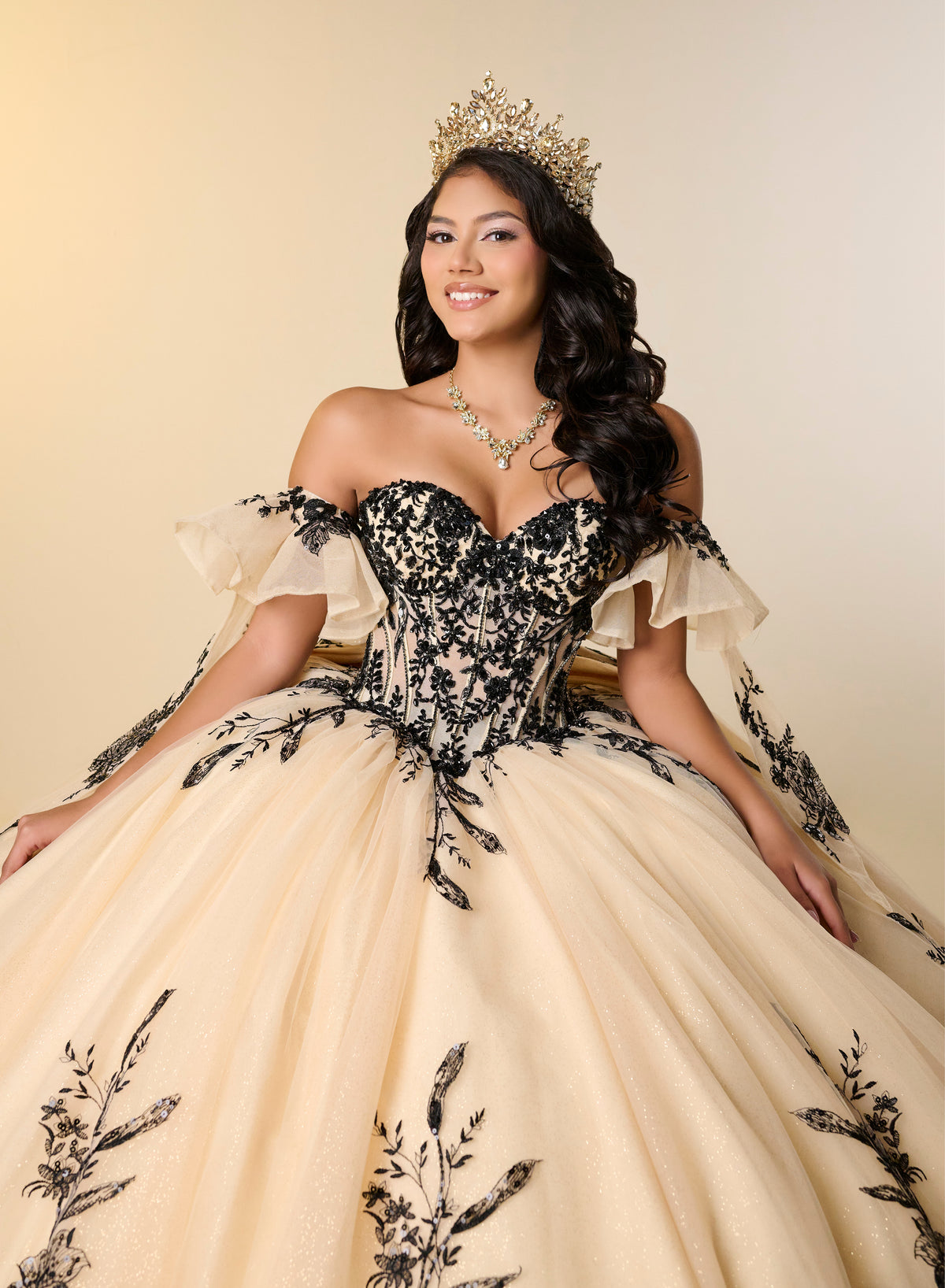 A woman wearing a voluminous off-the-shoulder quinceañera dress with a black lace corset bodice and beige tulle skirt with black floral embroidery, accessorized with a gold crown and necklace, posed against a warm beige studio background. / A strapless ball gown with a lace-up bodice and a voluminous tulle skirt featuring floral embroidery and a large back bow.