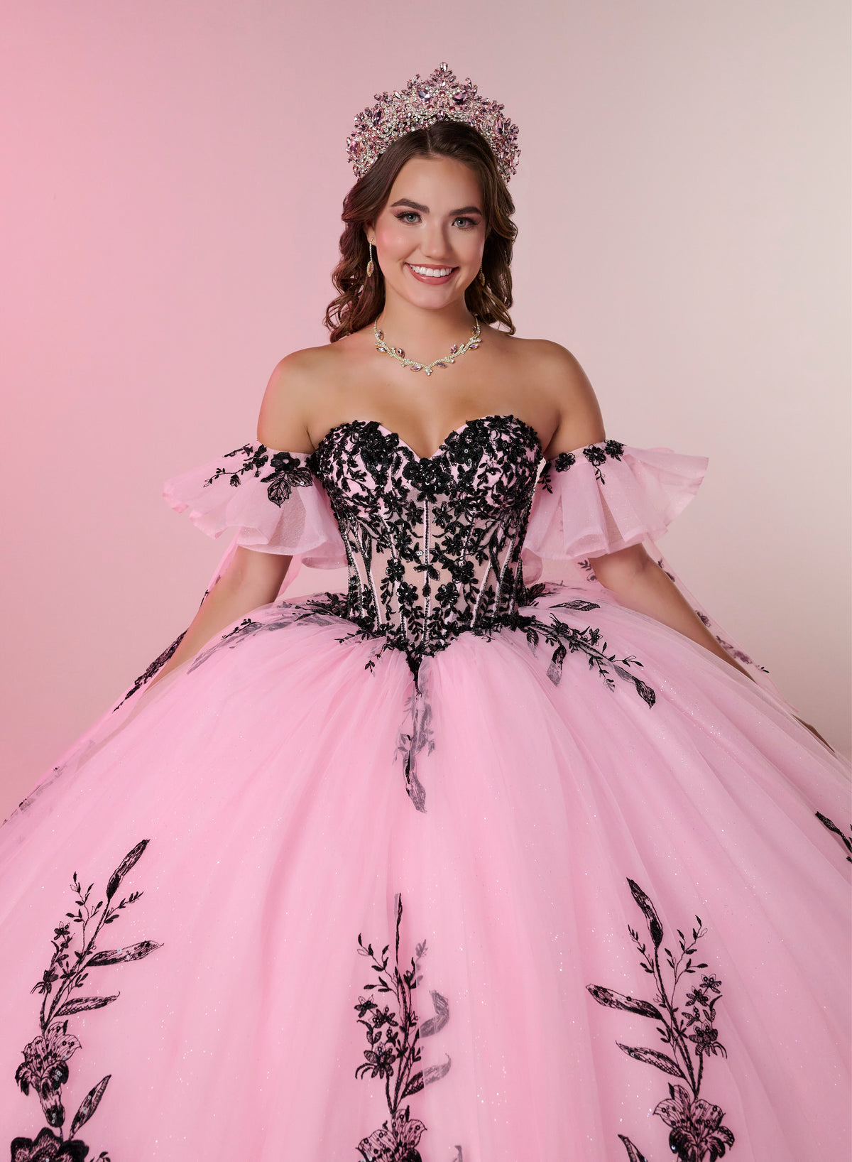 A smiling young woman wearing a voluminous pink ball gown with a black lace corset bodice, off-the-shoulder ruffled sleeves, and a matching pink tiara against a soft pink studio background. / A strapless ball gown with a lace-up bodice and a voluminous tulle skirt featuring floral embroidery and a large back bow.