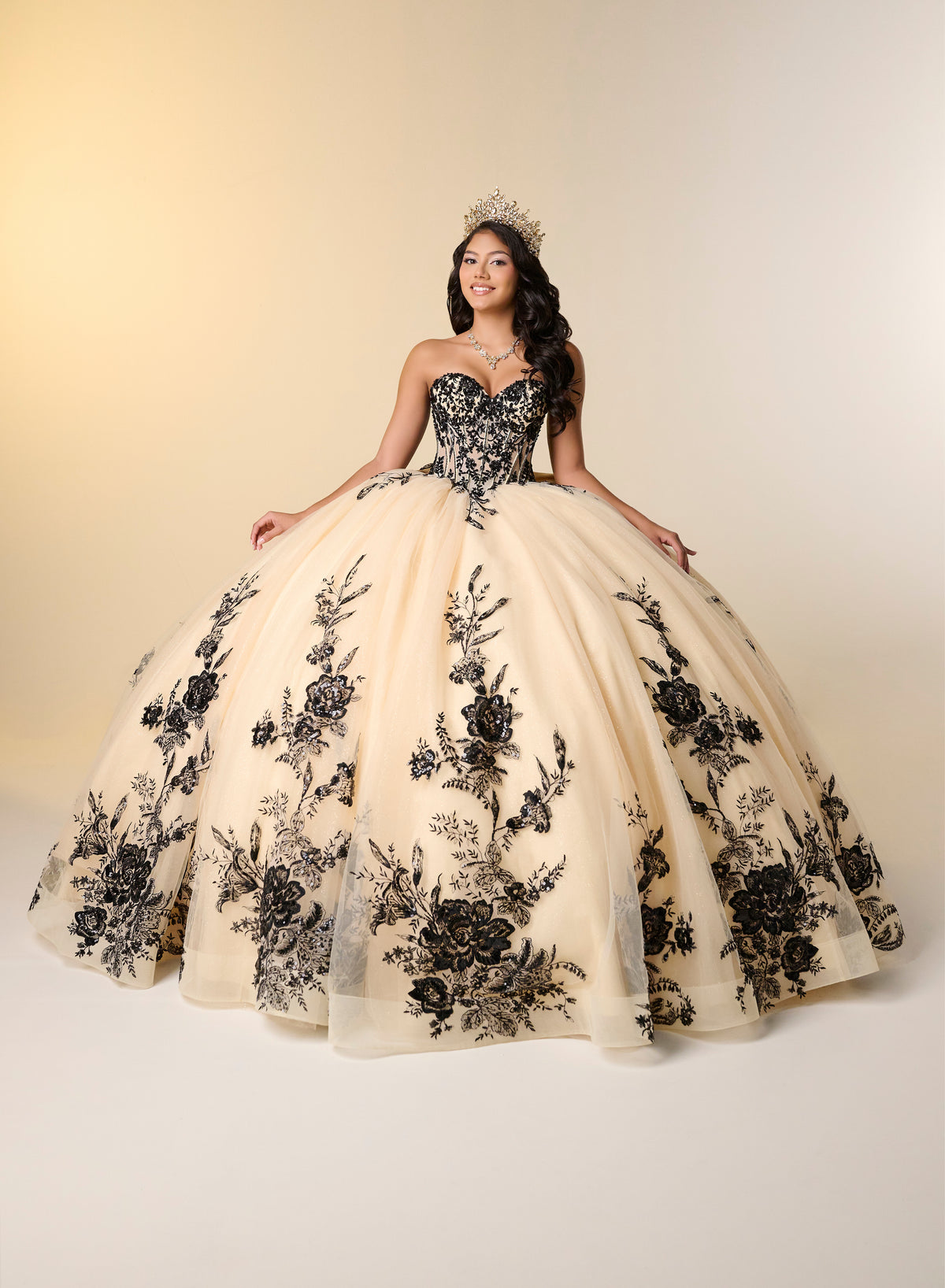 A woman wearing a strapless ball gown with a black floral lace bodice and a voluminous cream tulle skirt with black floral embroidery, posed against a warm beige studio background. / A strapless ball gown with a lace-up bodice and a voluminous tulle skirt featuring floral embroidery and a large back bow.