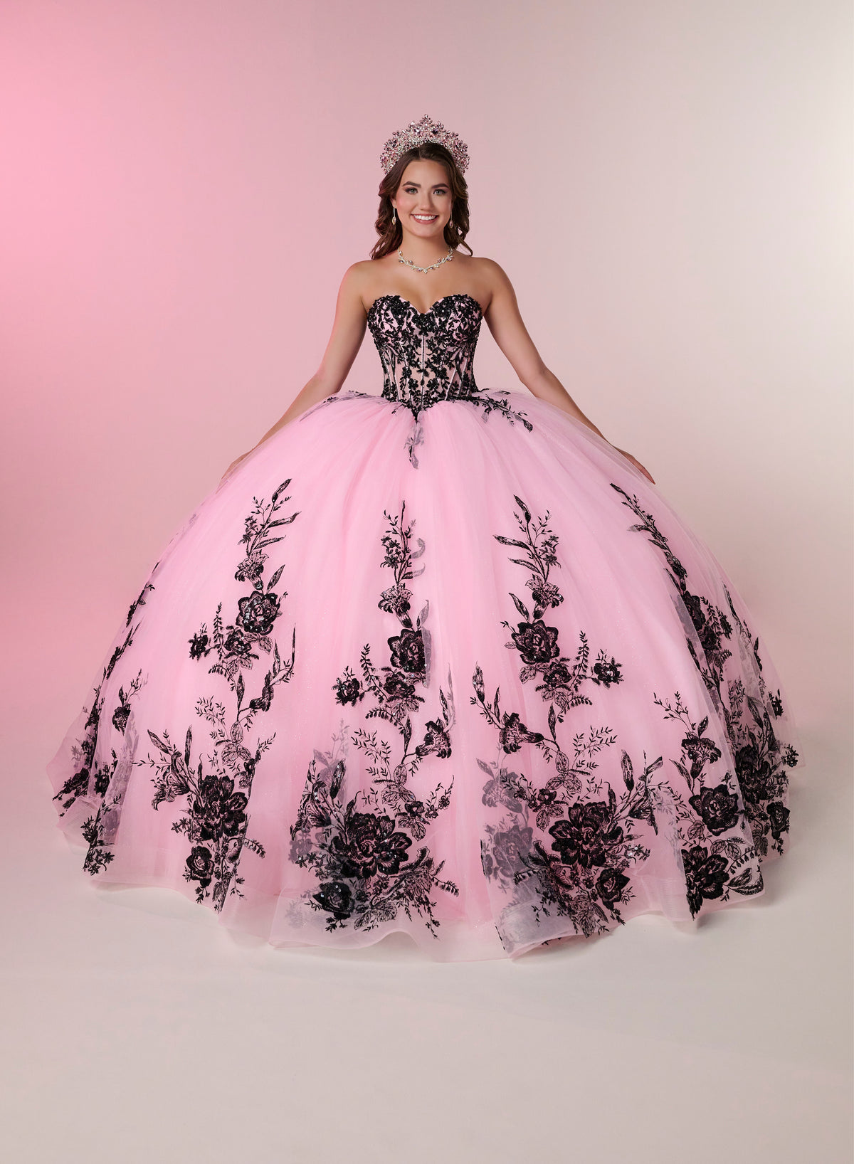 A young woman wearing a strapless pink ball gown with black floral lace applique and a tiara, posed against a soft pink studio backdrop. / A strapless ball gown with a lace-up bodice and a voluminous tulle skirt featuring floral embroidery and a large back bow.