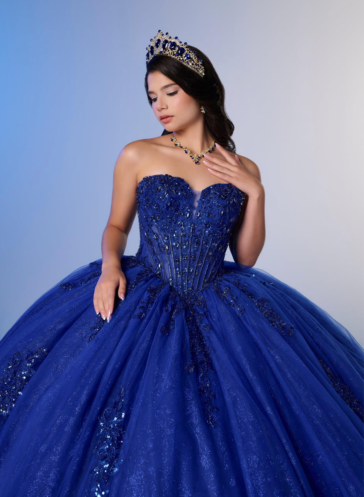 A woman wearing a royal blue strapless ball gown with beaded detailing, a matching tiara, and a necklace, posed against a blue-to-gray gradient studio background. / A strapless ball gown featuring a beaded corset bodice, a voluminous glitter-tulle skirt with sequin appliques, and a textured ruffle accent on the back.