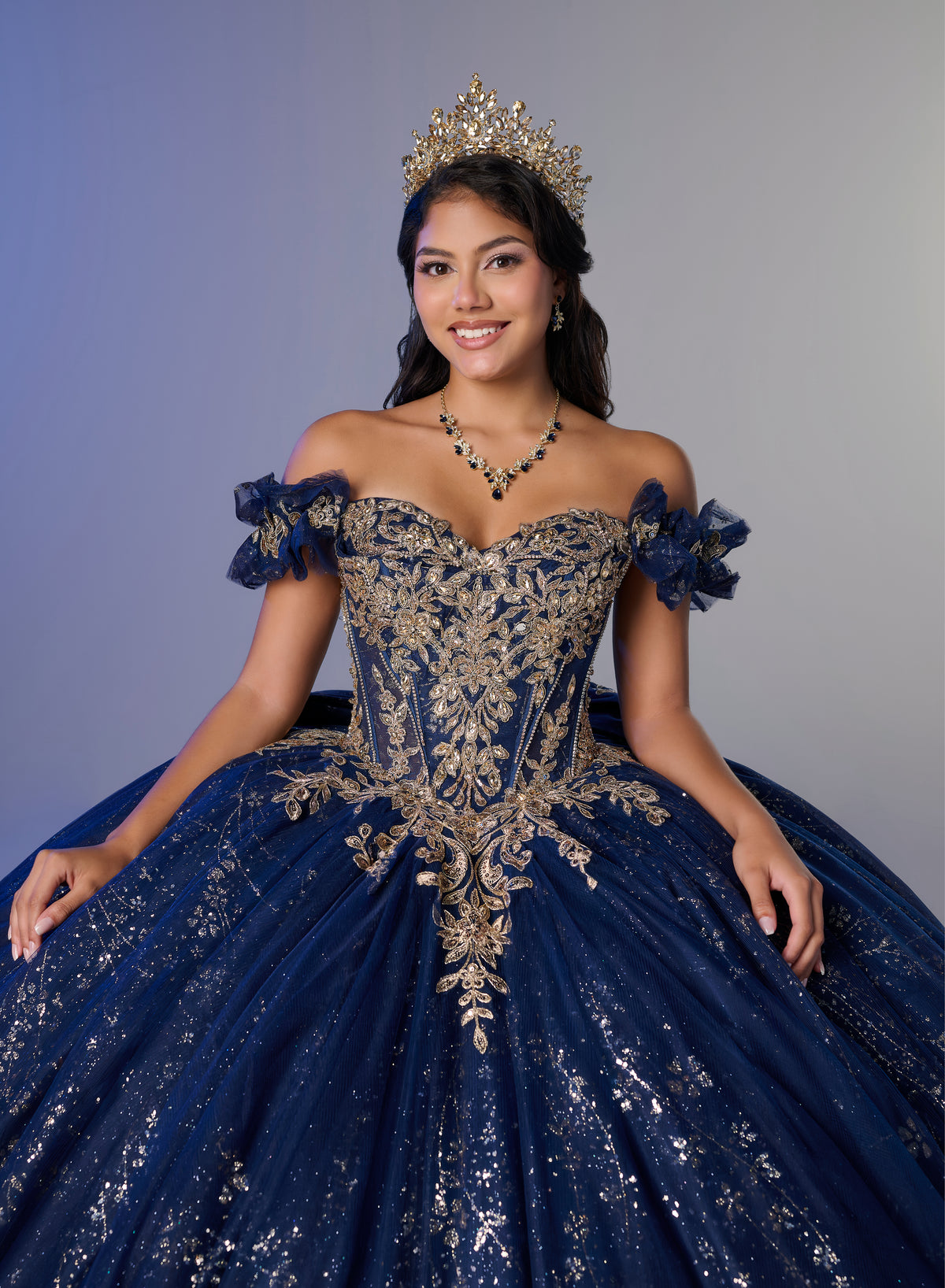 A young woman wearing an elaborate off-the-shoulder navy blue quinceañera dress with ornate gold embroidery, a matching tiara, and a necklace, posed against a solid gray studio background. / An off-the-shoulder ball gown with ruffled cap sleeves, a heavily embroidered metallic bodice, and a voluminous glitter-tulle skirt adorned with intricate baroque-style appliques and a matching oversized back bow.