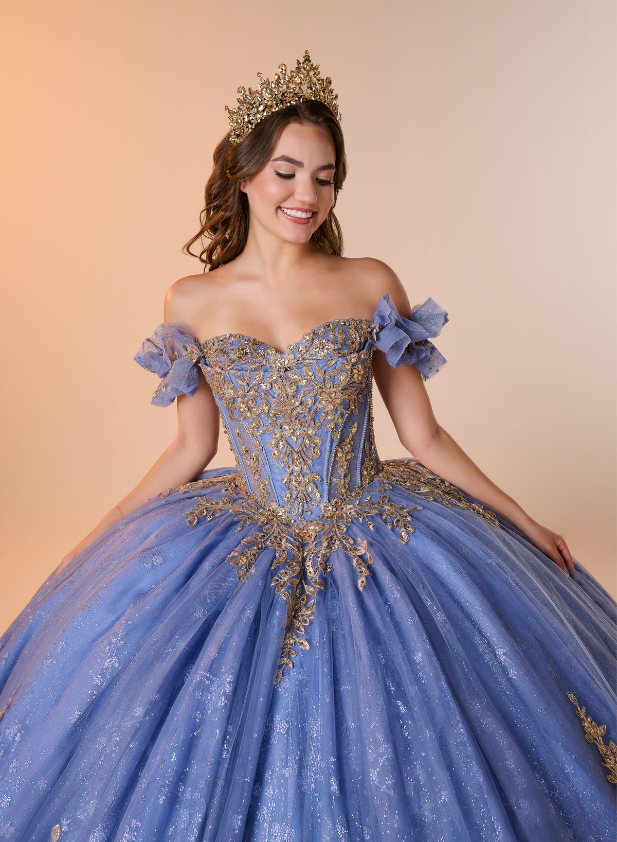 A woman wearing an off-the-shoulder light blue ball gown with ornate gold embroidery, a matching blue bow on the sleeve, and a gold tiara, posed against a warm beige studio background. / An off-the-shoulder ball gown with ruffled cap sleeves, a heavily embroidered metallic bodice, and a voluminous glitter-tulle skirt adorned with intricate baroque-style appliques and a matching oversized back bow.