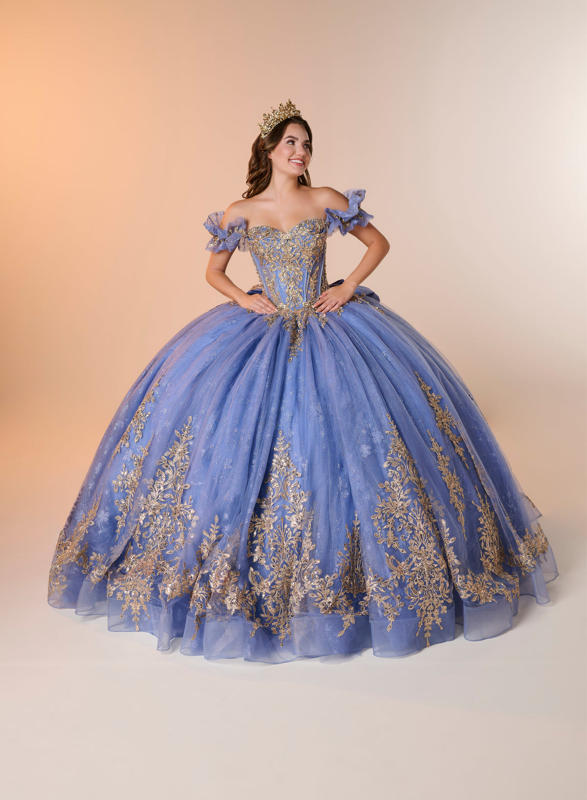 A woman modeling an off-the-shoulder blue quinceañera ball gown with gold floral embroidery and a matching gold crown, posed against a warm beige studio backdrop. / An off-the-shoulder ball gown with ruffled cap sleeves, a heavily embroidered metallic bodice, and a voluminous glitter-tulle skirt adorned with intricate baroque-style appliques and a matching oversized back bow.