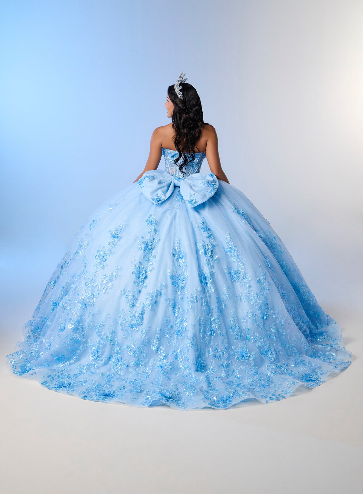 A model viewed from behind wearing a voluminous light blue ball gown with floral embroidery and a large bow at the back, set against a studio backdrop with a blue-to-white gradient. / A strapless sweetheart ball gown featuring a fully beaded corset bodice, a lace-up back, and a voluminous tulle skirt layered with shimmering sequin floral details and an oversized back bow, paired with a matching beaded choker necklace.