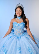 A young woman wearing a strapless light blue quinceañera dress with floral lace appliques, a matching choker, a silver tiara, and a full tulle skirt, posed against a light blue studio background. / A strapless sweetheart ball gown featuring a fully beaded corset bodice, a lace-up back, and a voluminous tulle skirt layered with shimmering sequin floral details and an oversized back bow, paired with a matching beaded choker necklace.
