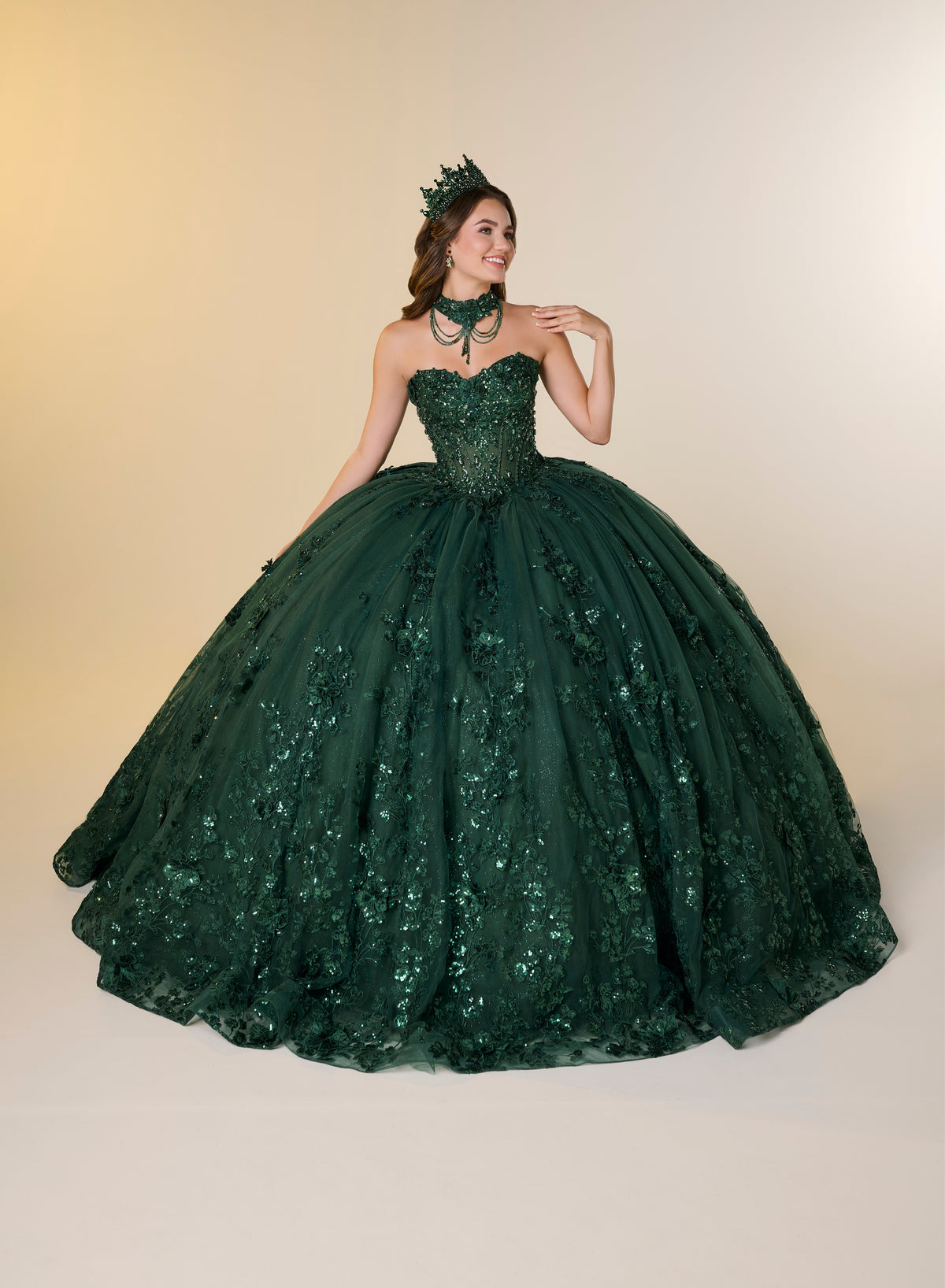 A woman modeling a strapless dark green ball gown with a sweetheart neckline, heavily embellished bodice, and a voluminous tulle skirt with floral lace and sequin details, against a warm beige studio background. / A strapless sweetheart ball gown featuring a fully beaded corset bodice, a lace-up back, and a voluminous tulle skirt layered with shimmering sequin floral details and an oversized back bow, paired with a matching beaded choker necklace.
