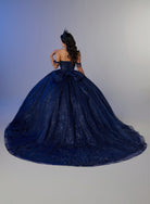 A woman photographed from behind wearing an elaborate off-the-shoulder ball gown in deep navy blue lace and tulle, with a large bow at the back of the bodice and a long train, against a soft gray studio background. / Ball gown with a lace-detailed back, corset lacing, and a large bow at the waist. The full skirt has a subtle sparkle and elegant embroidery.