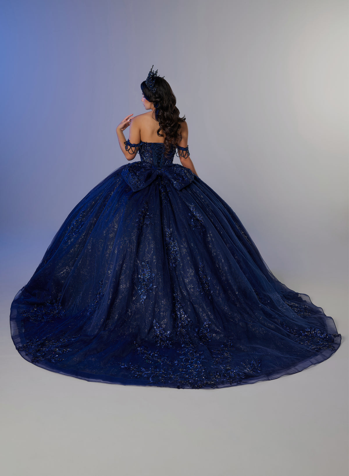 A woman photographed from behind wearing an elaborate off-the-shoulder ball gown in deep navy blue lace and tulle, with a large bow at the back of the bodice and a long train, against a soft gray studio background. / Ball gown with a lace-detailed back, corset lacing, and a large bow at the waist. The full skirt has a subtle sparkle and elegant embroidery.