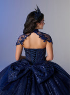 A woman shown from the back wearing an elaborate dark blue ball gown with lace shoulder details, a large bow at the back, and a matching tiara, against a soft gray studio background. / Ball gown with a lace-detailed back, corset lacing, and a large bow at the waist. The full skirt has a subtle sparkle and elegant embroidery.