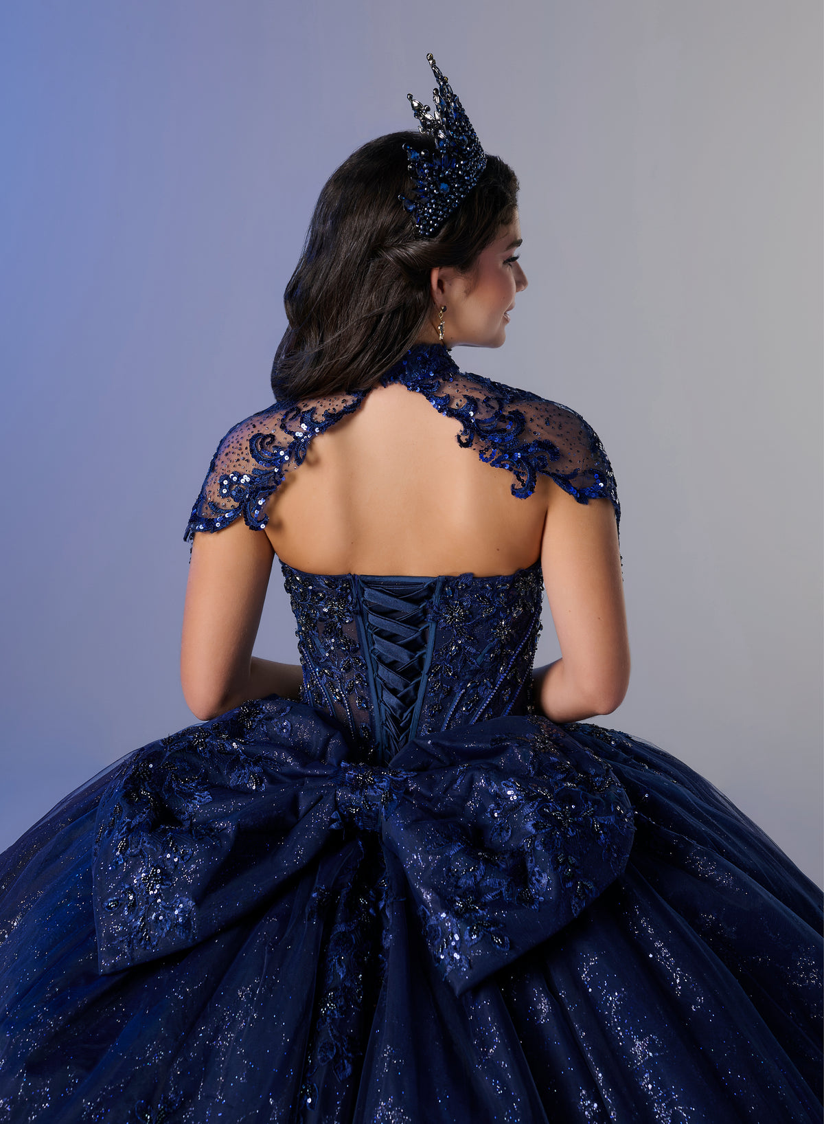 A woman shown from the back wearing an elaborate dark blue ball gown with lace shoulder details, a large bow at the back, and a matching tiara, against a soft gray studio background. / Ball gown with a lace-detailed back, corset lacing, and a large bow at the waist. The full skirt has a subtle sparkle and elegant embroidery.