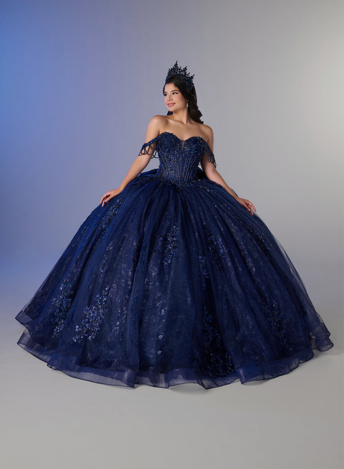 A woman wearing a voluminous navy blue ball gown with floral lace detailing and a matching tiara, posed against a neutral gray studio background. / Ball gown with a lace-detailed back, corset lacing, and a large bow at the waist. The full skirt has a subtle sparkle and elegant embroidery.