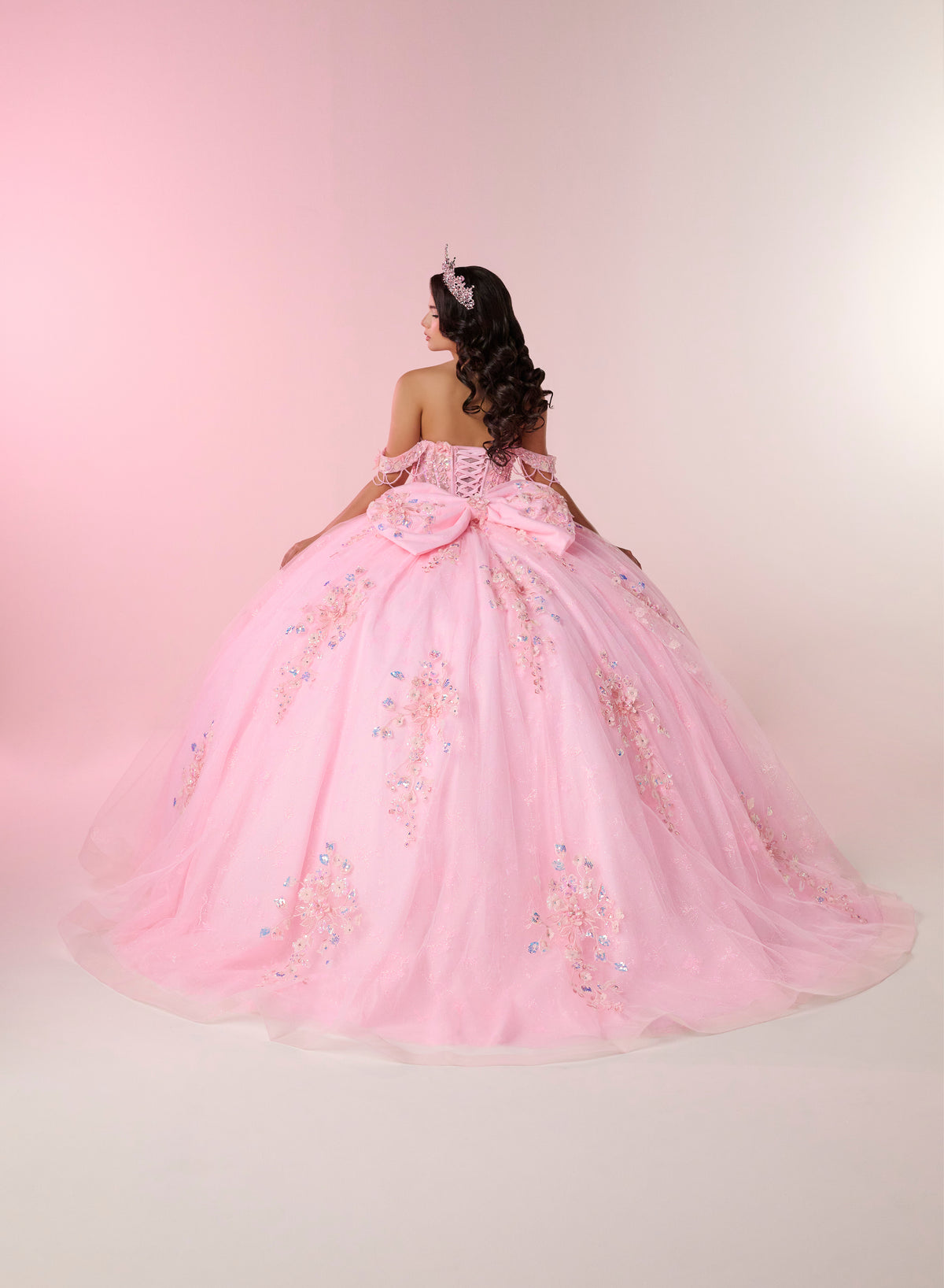 A young woman viewed from behind wearing a voluminous light pink ball gown with floral embroidery and a large bow at the back, posed against a soft pink studio backdrop. / An off-shoulder ball gown with a lace-up back, decorative bow, and floral embellishments on a full, layered skirt.