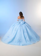 A woman viewed from behind wearing a voluminous light blue off-the-shoulder ball gown with floral embroidery and a large bow at the waist, photographed against a soft blue-to-white studio backdrop. / An off-shoulder ball gown with a lace-up back, decorative bow, and floral embellishments on a full, layered skirt.