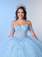 A smiling young woman wearing a sparkling off-the-shoulder light blue quinceañera dress with sequins and beadwork, a matching tiara, and a necklace, posed against a soft light blue studio background. / An off-shoulder ball gown with a lace-up back, decorative bow, and floral embellishments on a full, layered skirt.