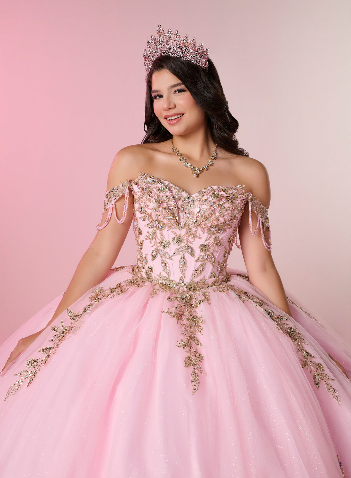 A young woman wearing a pink off-the-shoulder quinceañera dress with gold floral embroidery, a matching tiara, and a necklace, posing against a soft pink studio background. / Two-piece off-shoulder dress with a fitted, embellished bodice and a short, form-fitting skirt, featuring a sweetheart neckline, sparkling embroidered details, detachable arm drapes, and a full tulle skirt with a sweeping train.