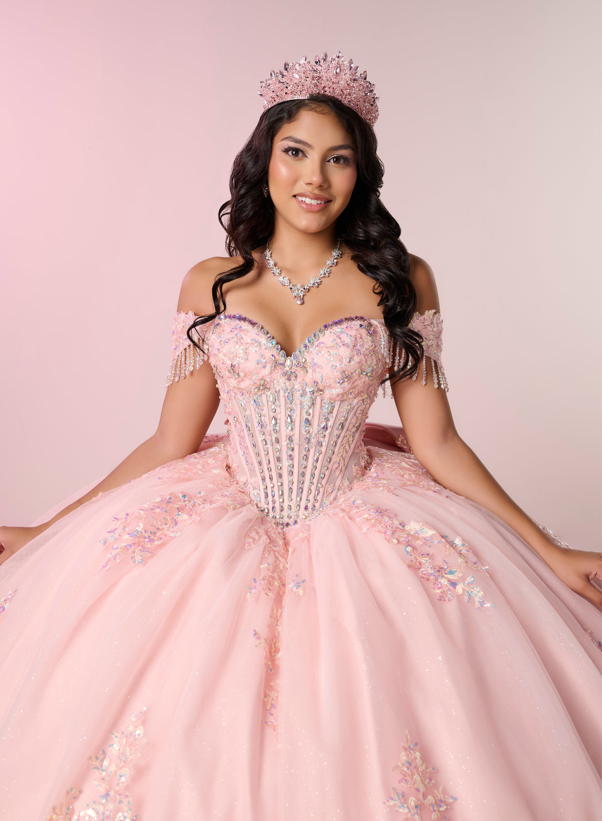 A woman wearing a voluminous light pink ball gown with a corset-style bodice, off-the-shoulder sleeves, and sparkling embellishments, posed against a soft pink studio background. She is also wearing a pink tiara and a matching necklace. / Luxurious off-the-shoulder ball gown with intricate detailing and a statement bow at the back.