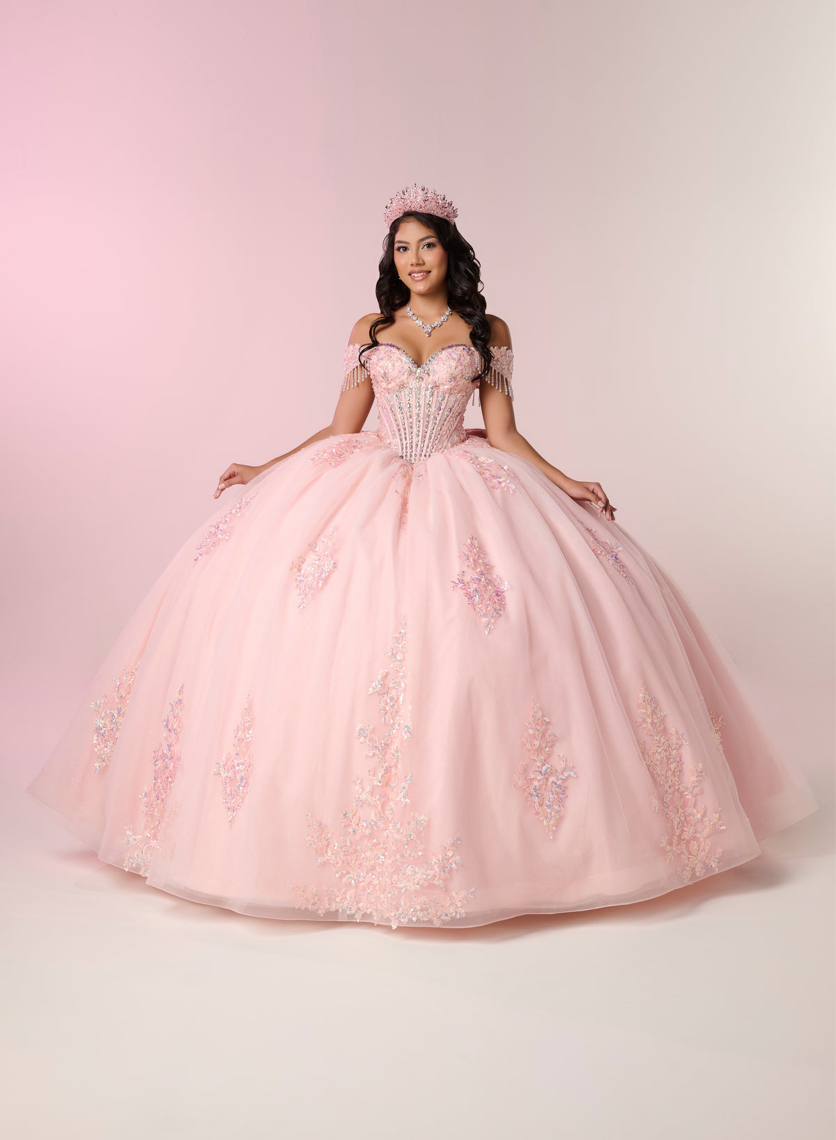 A young woman wearing a voluminous light pink ball gown with floral embroidery and a matching pink tiara, posed against a soft pink studio backdrop. / Luxurious off-the-shoulder ball gown with intricate detailing and a statement bow at the back.