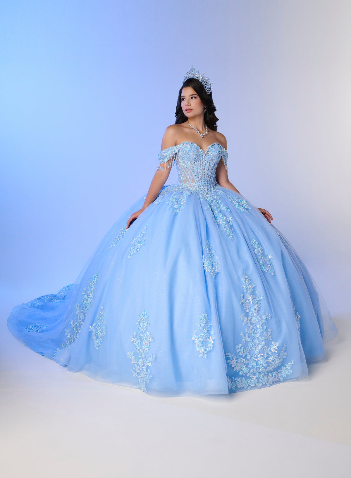 A woman wearing a voluminous light blue off-the-shoulder ball gown with floral embroidery and a matching tiara, posed against a soft blue-to-white studio backdrop. / Luxurious off-the-shoulder ball gown with intricate detailing and a statement bow at the back.
