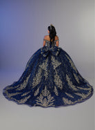 A woman viewed from behind wearing an elaborate off-the-shoulder ball gown with a very full skirt and large bow at the back. The dress is dark blue with ornate metallic gold embroidery, set against a plain studio background. / Elegant off-shoulder ball gown featuring intricate embroidered detailing, a structured bodice, and a voluminous skirt with a dramatic statement bow at the back.