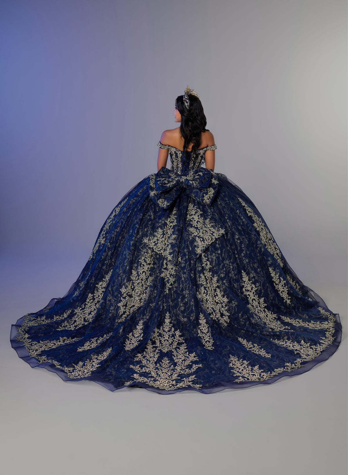 A woman viewed from behind wearing an elaborate off-the-shoulder ball gown with a very full skirt and large bow at the back. The dress is dark blue with ornate metallic gold embroidery, set against a plain studio background. / Elegant off-shoulder ball gown featuring intricate embroidered detailing, a structured bodice, and a voluminous skirt with a dramatic statement bow at the back.