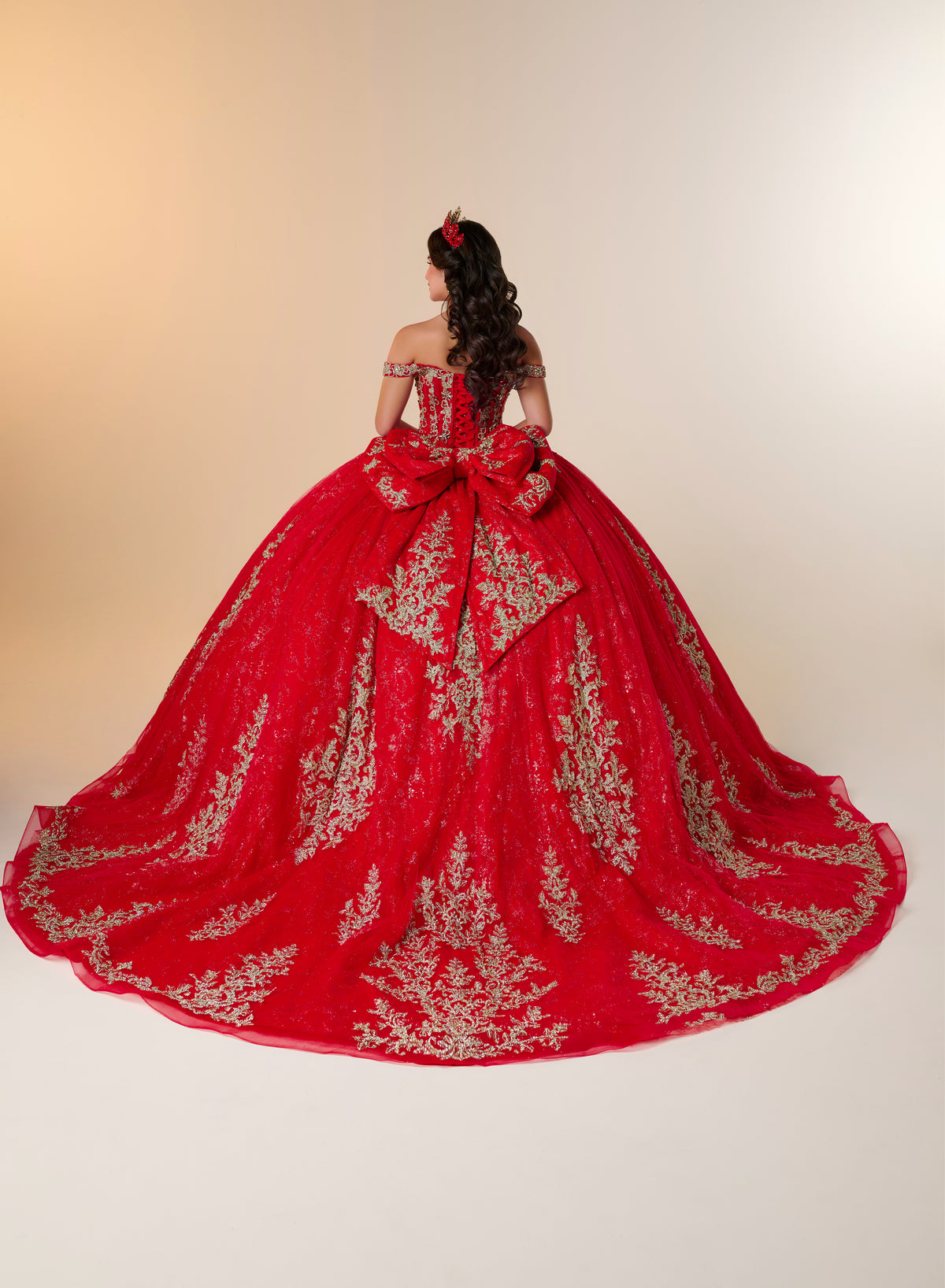 A red off-the-shoulder ball gown with intricate gold embroidery, modeled from behind to show the large bow at the back of the bodice and the voluminous train against a plain beige studio background. / Elegant off-shoulder ball gown featuring intricate embroidered detailing, a structured bodice, and a voluminous skirt with a dramatic statement bow at the back.