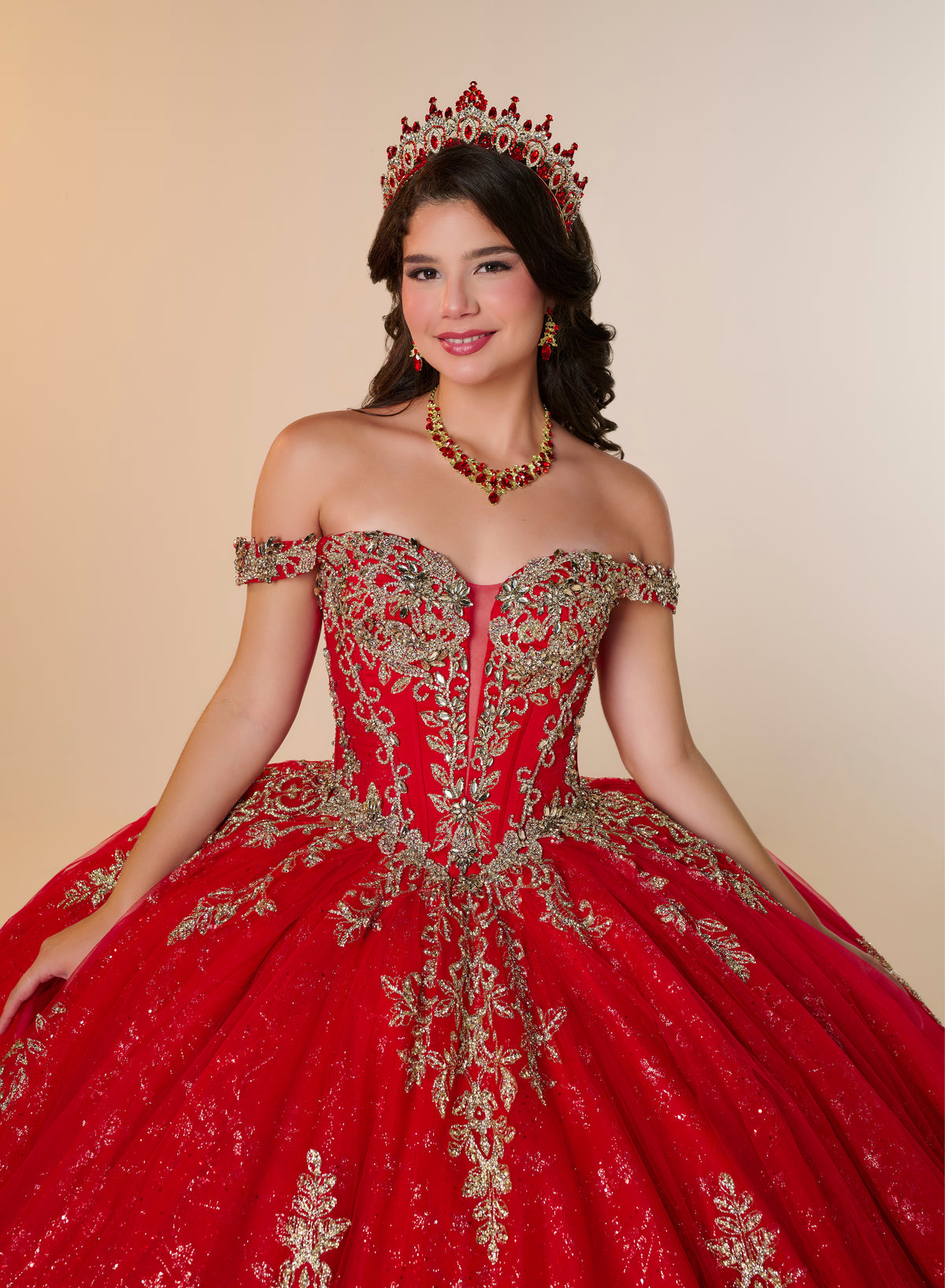 A young woman modeling a red off-the-shoulder ball gown with extensive gold embroidery, wearing a matching red and gold tiara and necklace against a solid beige background. / Elegant off-shoulder ball gown featuring intricate embroidered detailing, a structured bodice, and a voluminous skirt with a dramatic statement bow at the back.