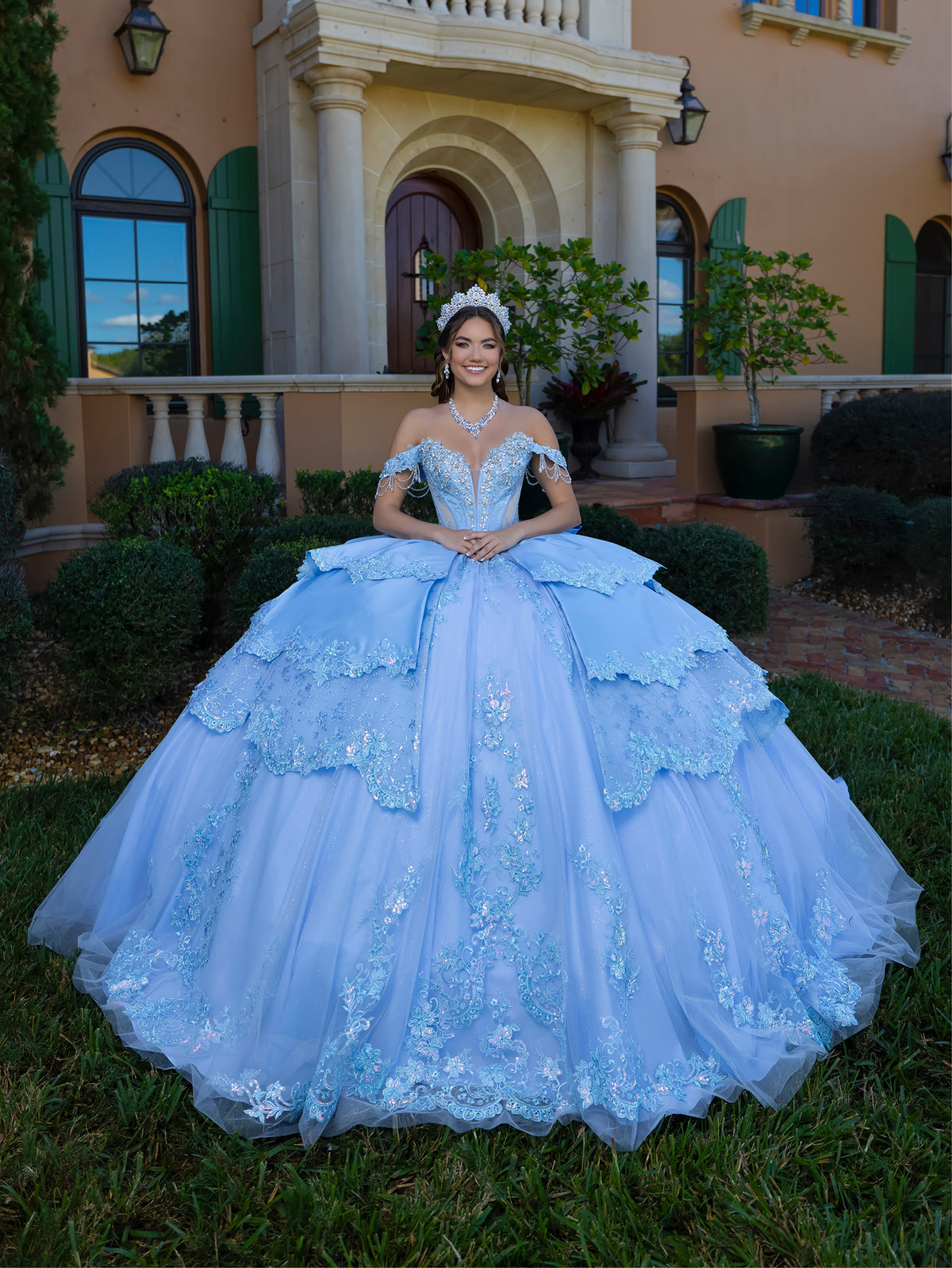 This gown features a fitted, embellished bodice with off-the-shoulder sleeves and a full tiered ball-gown skirt. Intricate appliqués decorate the layers, and a long flowing train with a large back bow completes the princess-style look.
