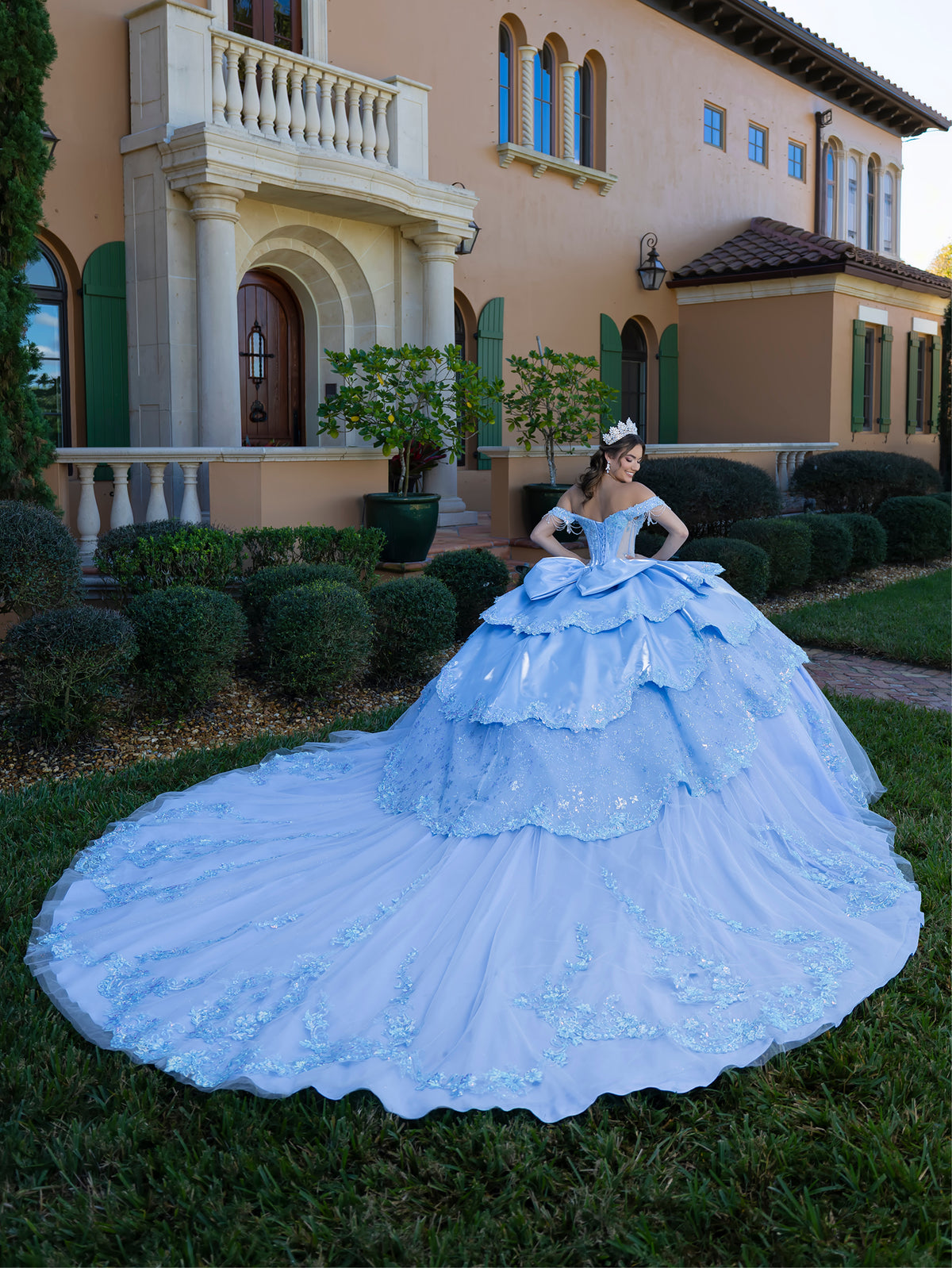 This gown features a fitted, embellished bodice with off-the-shoulder sleeves and a full tiered ball-gown skirt. Intricate appliqués decorate the layers, and a long flowing train with a large back bow completes the princess-style look.