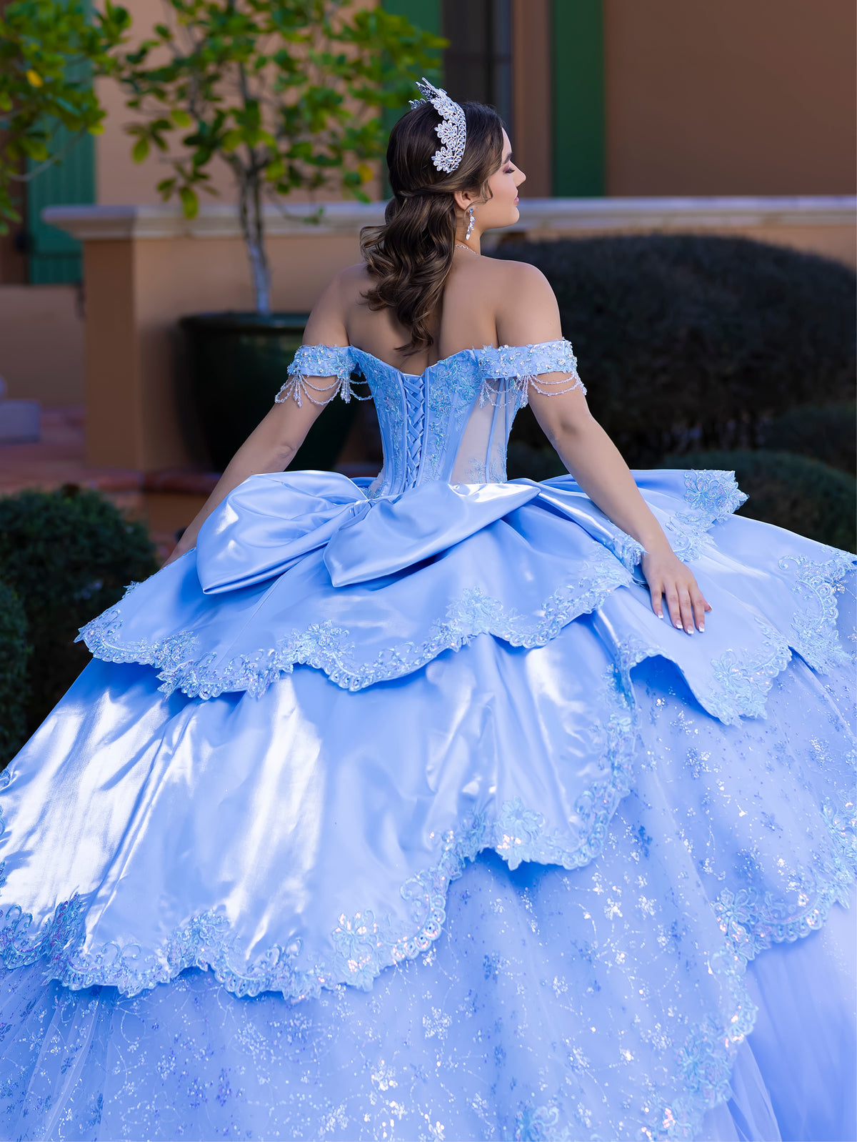 This gown features a fitted, embellished bodice with off-the-shoulder sleeves and a full tiered ball-gown skirt. Intricate appliqués decorate the layers, and a long flowing train with a large back bow completes the princess-style look.