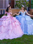 Two young women wearing elaborate off-the-shoulder quinceañera ballgowns stand on grass outdoors, holding hands. One gown is light pink and the other is light blue, both with full tiered ruffles, embroidered bodices, and matching tiara headpieces. / This gown features a fitted, embellished bodice with off-the-shoulder sleeves and a full tiered ball-gown skirt. Intricate appliqués decorate the layers, and a long flowing train with a large back bow completes the princess-style look.