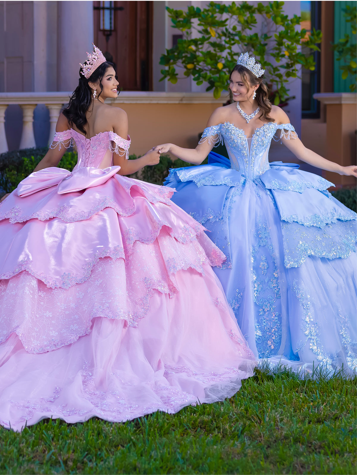 Two young women wearing elaborate off-the-shoulder quinceañera ballgowns stand on grass outdoors, holding hands. One gown is light pink and the other is light blue, both with full tiered ruffles, embroidered bodices, and matching tiara headpieces. / This gown features a fitted, embellished bodice with off-the-shoulder sleeves and a full tiered ball-gown skirt. Intricate appliqués decorate the layers, and a long flowing train with a large back bow completes the princess-style look.