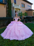 This gown features a fitted, embellished bodice with off-the-shoulder sleeves and a full tiered ball-gown skirt. Intricate appliqués decorate the layers, and a long flowing train with a large back bow completes the princess-style look.