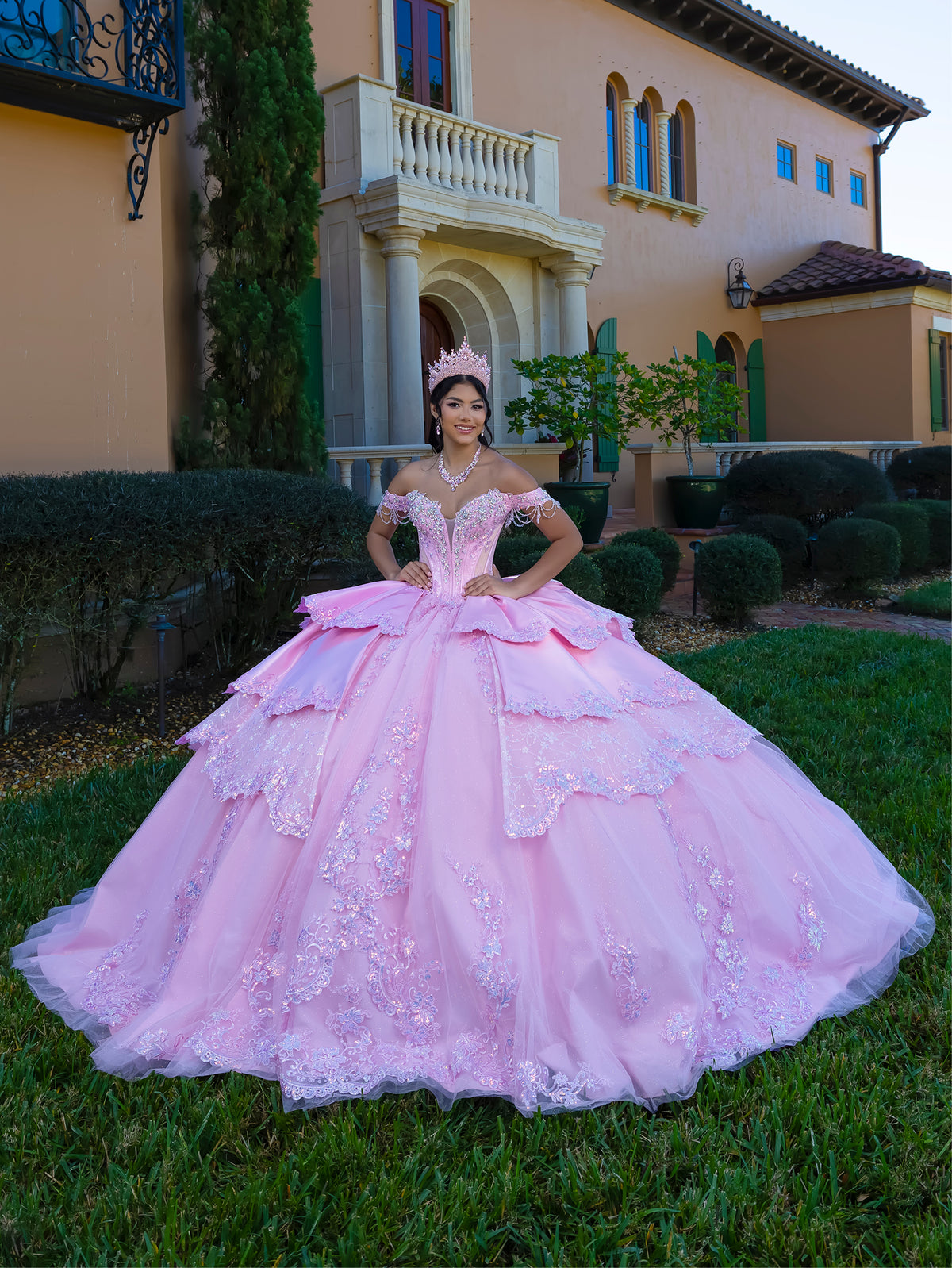 This gown features a fitted, embellished bodice with off-the-shoulder sleeves and a full tiered ball-gown skirt. Intricate appliqués decorate the layers, and a long flowing train with a large back bow completes the princess-style look.